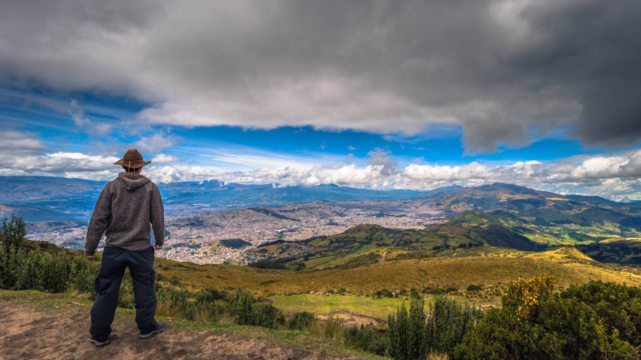 Persona de pie en la ladera de una colina que domina un vasto y poblado valle con nubes dispersas en el cielo.