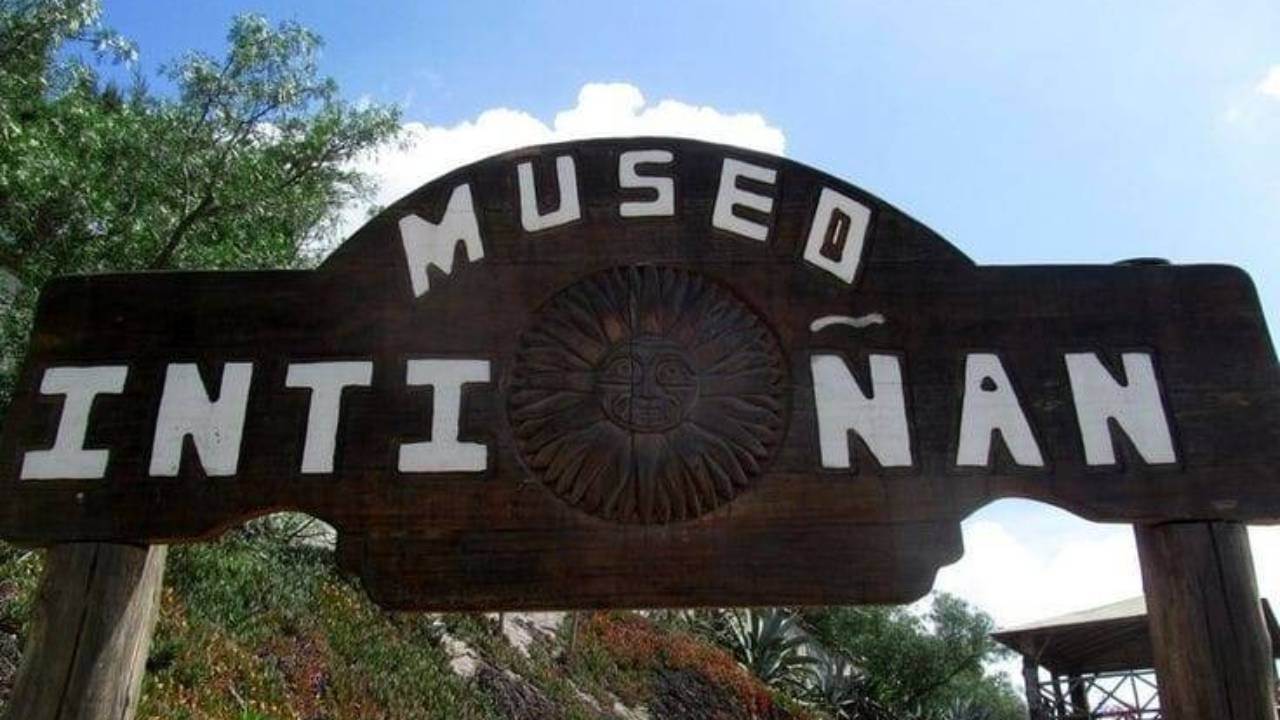 Un cartel de madera con el texto "Museo Intiñan" y una cara de sol tallada en el centro. El fondo incluye vegetación y una estructura.