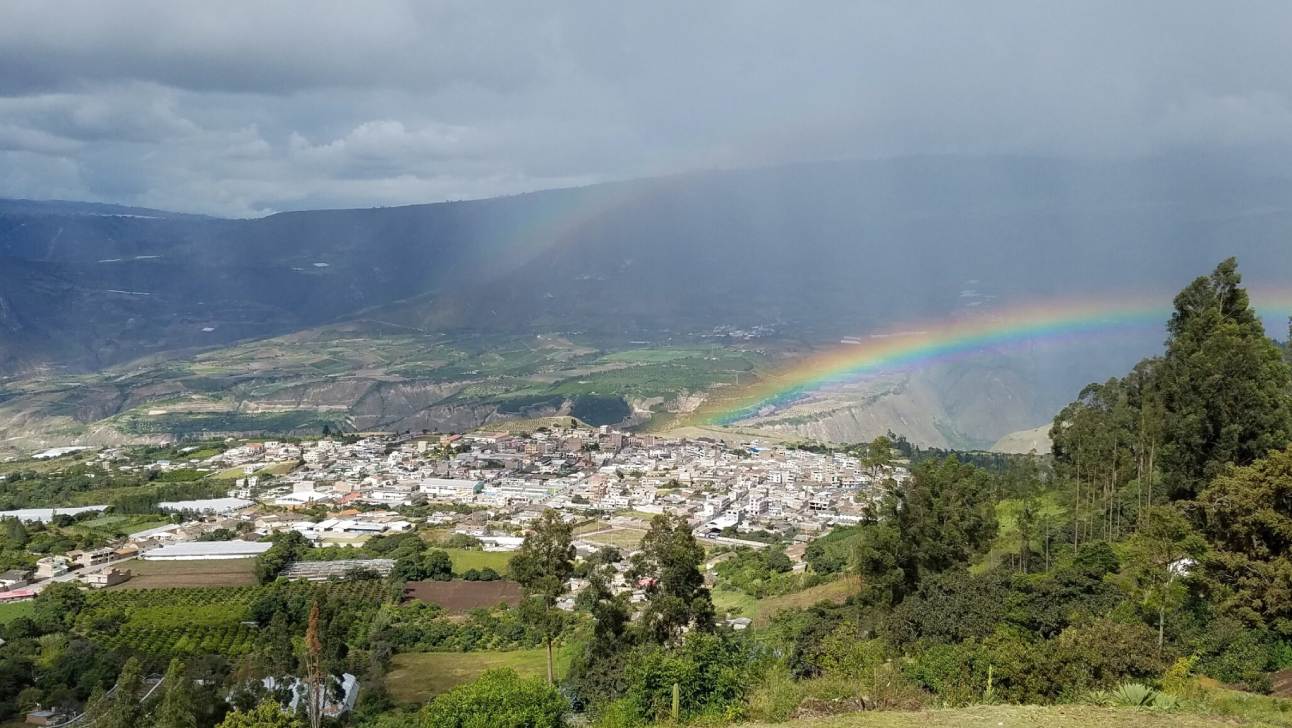 Una ciudad está situada en un valle verde con un arco iris atravesándolo; montañas y un cielo nublado están al fondo.