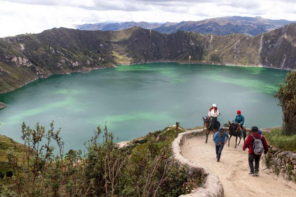 Dos personas van a caballo y otras dos caminan por un sendero de tierra desde el que se divisa un gran lago verde en un cráter volcánico.
