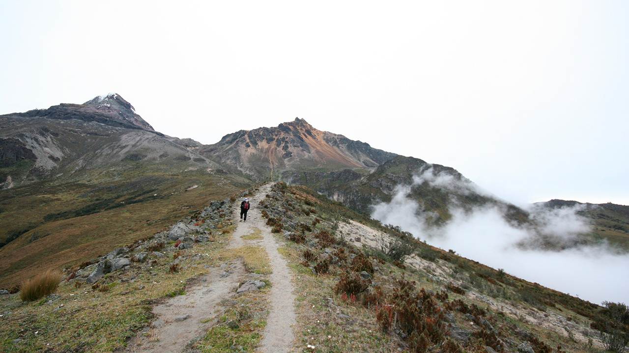 Una persona camina por un sendero de montaña con nubes al fondo.
