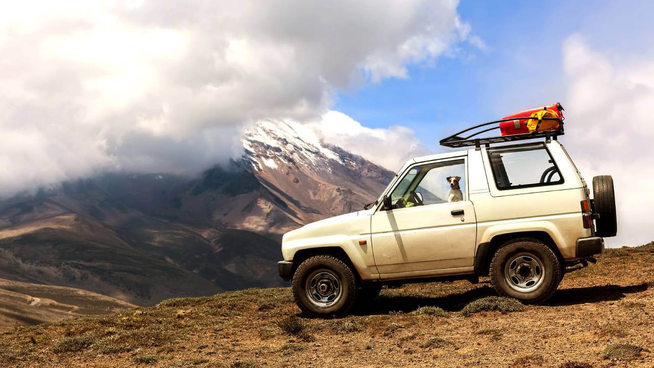 Un todoterreno blanco estacionado en un terreno montañoso con un pico cubierto de nieve al fondo bajo un cielo azul nublado.