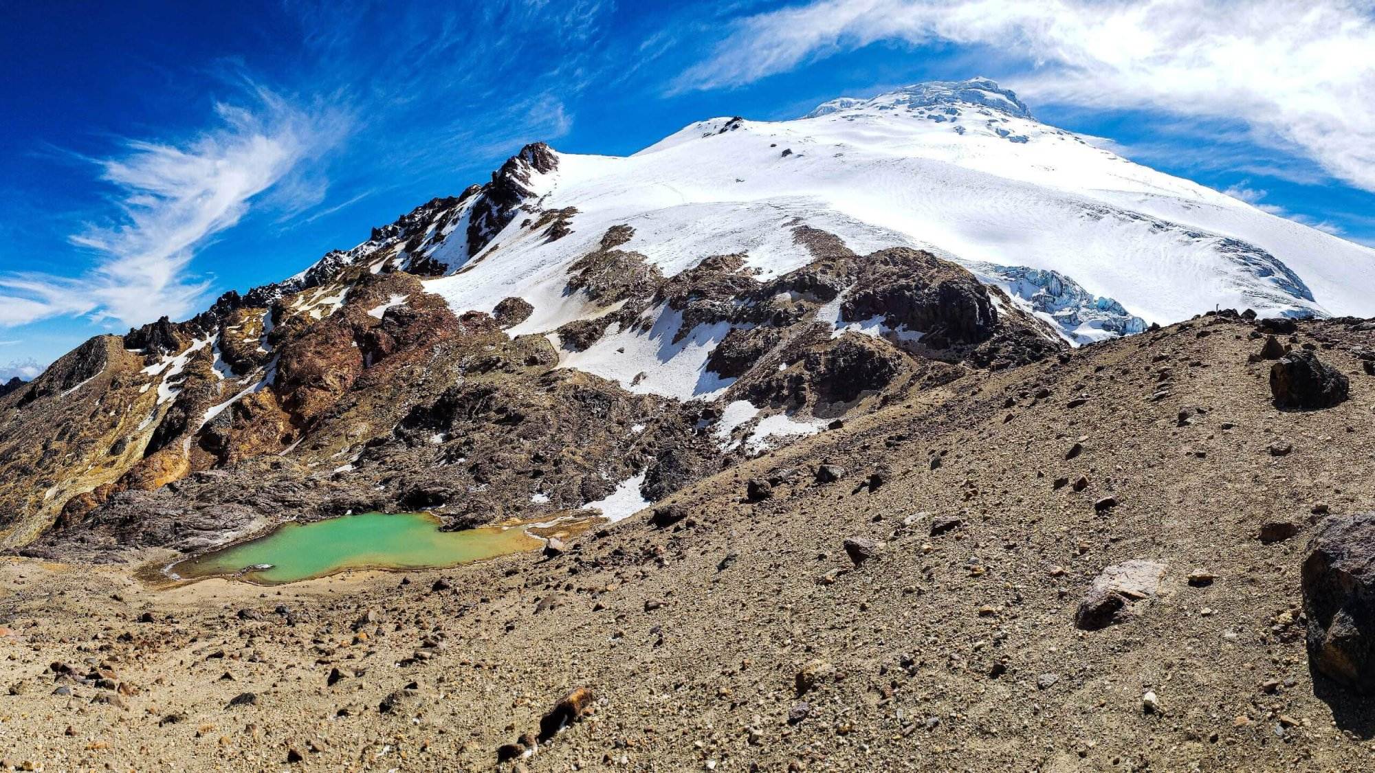 Monta&ntilde;a cubierta de nieve con terreno rocoso en primer plano y un peque&ntilde;o lago turquesa bajo un cielo azul brillante.