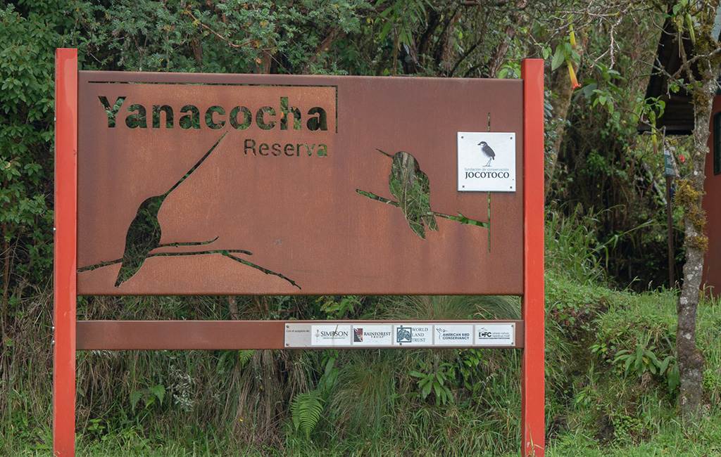 Letrero de metal rústico para la Reserva Yanacocha con recortes de aves y un logotipo de Jocotoco, sobre un fondo de vegetación densa.